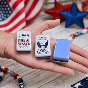 May include: Three rectangular mahjong tiles with rounded corners. One tile features the text JOKER USA Fourth of July with a flag pattern. Another tile displays the text WHITE DRAGON with a bald eagle graphic. The third tile is solid light blue.
