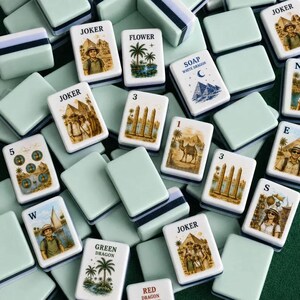 K&ouml;nnte beinhalten: Eine Sammlung rechteckiger Mahjong-Spielsteine mit hellgr&uuml;nen R&uuml;ckseiten und wei&szlig;en Vorderseiten, die verschiedene Illustrationen wie Pyramiden, Palmen, Kamele und Personen zeigen. Einige Steine tragen Aufschriften wie JOKER, FLOWER, SOAP und WHITE DRAGON.