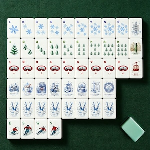 Pu&ograve; includere: Un set di tessere Mahjong bianche con illustrazioni a tema invernale, tra cui fiocchi di neve, alberi di pino, occhiali da sci e una funivia rossa. Le tessere sono disposte su una superficie verde, con una tessera verde abbinata.