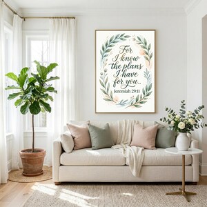 Op de afbeelding: Een rechthoekige wandkunstprint met een witte achtergrond, versierd met een krans van groene bladeren en zachte aquarelaccenten. De tekst luidt: For I know the plans I have for you.. Jeremiah 29:11 in een donker, elegant handschriftlettertype.