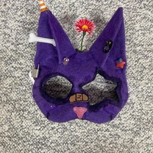 Op de afbeelding: Een paars vilten kattenmasker met diverse versieringen. Het heeft een feesthoedje, een bot, een rode bloem en diverse kleine decoratieve elementen. Het masker heeft ooggaten en een neus, met een pleister en een roze neus.