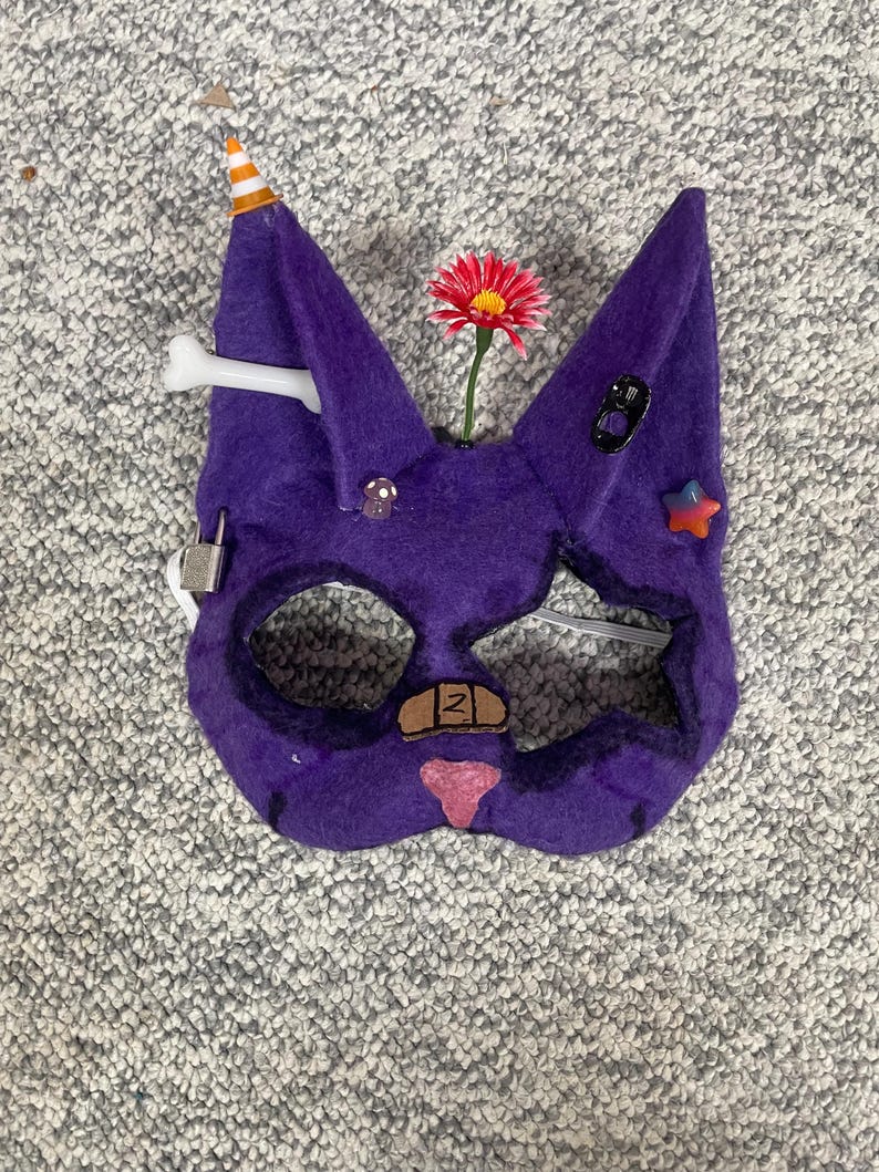 Op de afbeelding: Een paars vilten kattenmasker met diverse versieringen. Het heeft een feesthoedje, een bot, een rode bloem en diverse kleine decoratieve elementen. Het masker heeft ooggaten en een neus, met een pleister en een roze neus.
