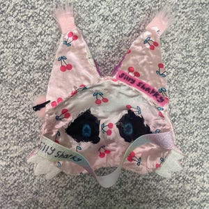 Op de afbeelding: Een roze slaapmasker met een kersenpatroon en de tekst "Silly Sharks". Het masker heeft zwarte ooggaten met blauwe accenten, een witte band en roze afwerking. Het masker is gemaakt van een zacht, zijdeachtig materiaal.