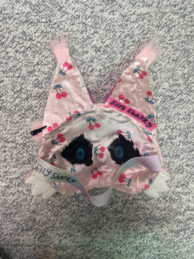Op de afbeelding: Een roze slaapmasker met een kersenpatroon en de tekst "Silly Sharks". Het masker heeft zwarte ooggaten met blauwe accenten, een witte band en roze afwerking. Het masker is gemaakt van een zacht, zijdeachtig materiaal.
