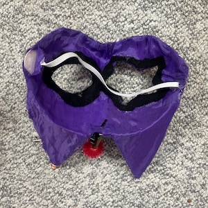 Op de afbeelding: Een paars kostuummasker met zwarte ooggaten en een witte elastische band. Het masker heeft een rode pompon aan de onderkant bevestigd. Het masker is gemaakt van een glanzende stof.