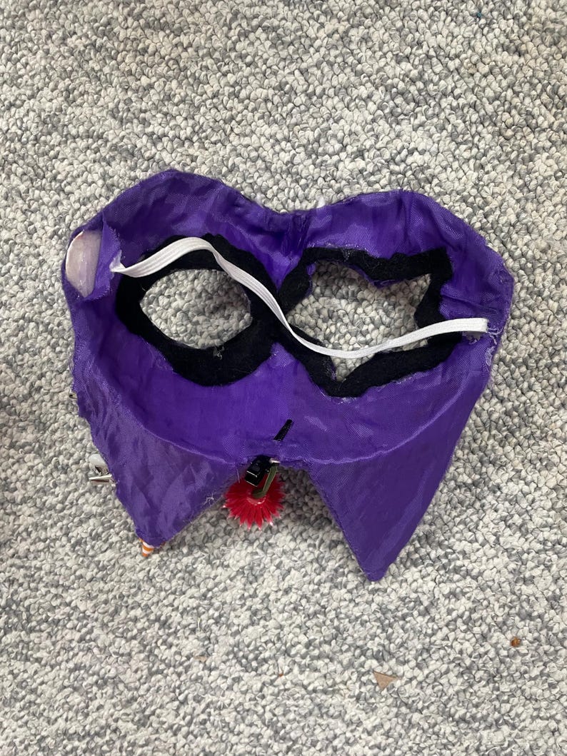 Op de afbeelding: Een paars kostuummasker met zwarte ooggaten en een witte elastische band. Het masker heeft een rode pompon aan de onderkant bevestigd. Het masker is gemaakt van een glanzende stof.