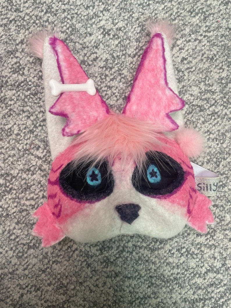 Op de afbeelding: Een roze en wit pluche masker in de vorm van een cartoon vossengezicht. Het masker heeft grote zwarte ogen met blauwe stervormige pupillen, roze oren en een wit botvormig detail. Het heeft pluizig roze bont rond de ogen en een klein label met de tekst "Silly".