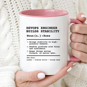 May include: A white ceramic mug with a pink interior and handle, featuring black printed text that defines a DevOps Engineer as someone who builds stability, lists three professional traits, and includes a See Also section with related terms.