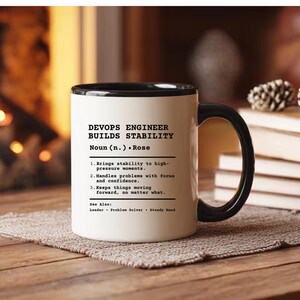 May include: A white ceramic mug with a black interior and handle, featuring black printed text that defines a DevOps Engineer as someone who builds stability, handles high-pressure moments, and keeps things moving forward with focus.