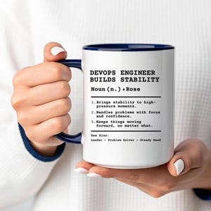 May include: A white ceramic mug with a dark blue handle and rim, featuring black printed text that defines a DevOps Engineer as someone who brings stability to high-pressure moments, handles problems with focus, and keeps things moving forward.