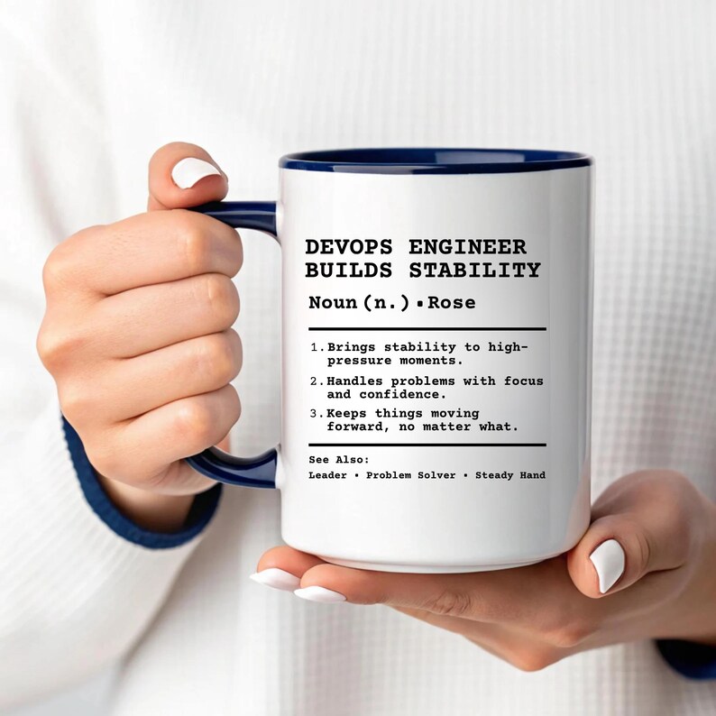 May include: A white ceramic mug with a dark blue handle and rim, featuring black printed text that defines a DevOps Engineer as someone who brings stability to high-pressure moments, handles problems with focus, and keeps things moving forward.