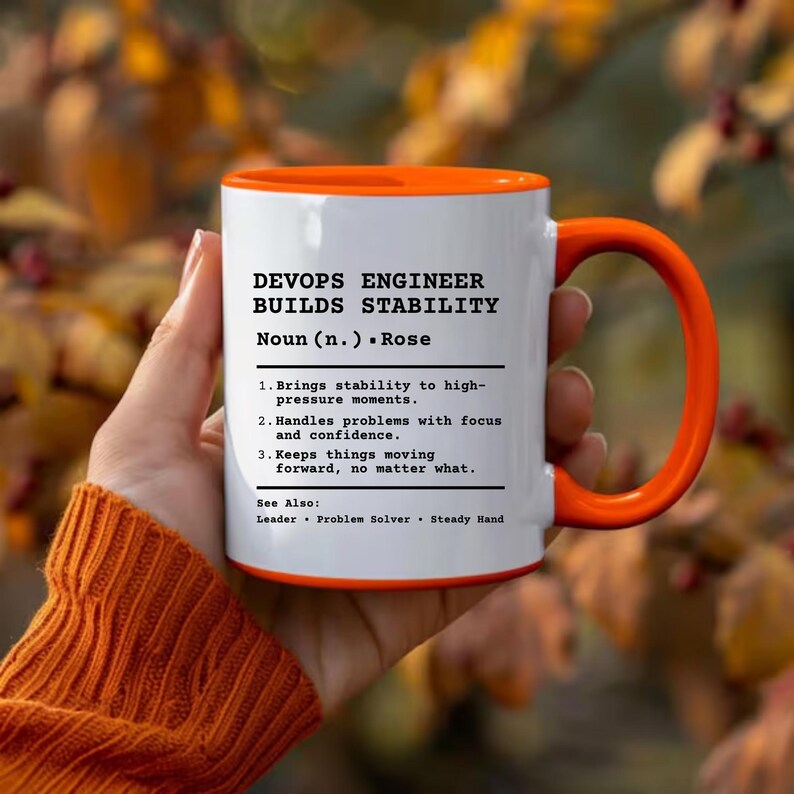 May include: A white ceramic mug with a bright orange interior and handle. The front features black text defining a DevOps Engineer as someone who brings stability to high-pressure moments, handles problems with focus, and keeps things moving forward.