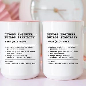 May include: Two white ceramic mugs with light pink handles and interior rims. The mugs feature black printed text that reads: DEVOPS ENGINEER BUILDS STABILITY, Noun (n.) Rose, 1. Brings stability to high-pressure moments, 2. Handles problems with focus and confidence, 3. Keeps things moving forward, no matter what. See Also: Leader, Problem Solver, Steady Hand.