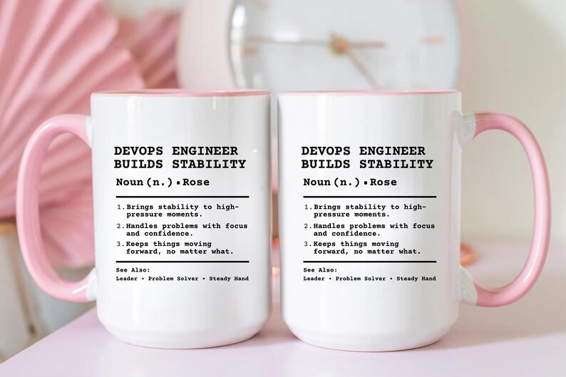 May include: Two white ceramic mugs with light pink handles and interior rims. The mugs feature black printed text that reads: DEVOPS ENGINEER BUILDS STABILITY, Noun (n.) Rose, 1. Brings stability to high-pressure moments, 2. Handles problems with focus and confidence, 3. Keeps things moving forward, no matter what. See Also: Leader, Problem Solver, Steady Hand.