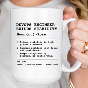 May include: A white ceramic mug featuring black printed text that defines a DevOps Engineer as someone who builds stability. The text includes a dictionary-style entry with three numbered points and a 'See Also' section listing Leader, Problem Solver, and Steady Hand.