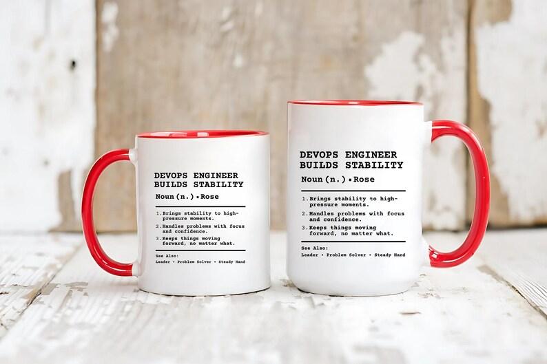 May include: Two white ceramic mugs with bright red handles and interiors. Each mug features black printed text that defines a DevOps Engineer as someone who builds stability, including the name Rose and three numbered points describing professional traits.