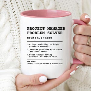 May include: A white ceramic mug with a pink interior and handle, featuring black printed text that defines a Project Manager as a problem solver. The text includes a dictionary-style entry with three numbered points and a see also section.