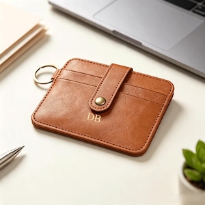 May include: A tan rectangular card holder with a textured finish, featuring a snap button closure, a metal key ring attached to the side, and the letters DB embossed in gold on the front surface. The item has visible stitching along the edges.