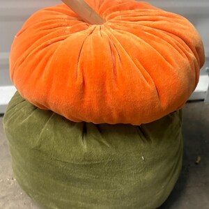 May include: A stack of two round, plush, velvet-textured decorative pillows shaped like pumpkins. The top pillow is bright orange with a light brown wooden stem, and the bottom pillow is olive green. Both pillows feature gathered fabric.