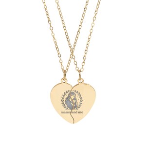 Peut inclure: Un collier doré avec deux pendentifs en forme de cœur qui s'emboîtent. Chaque pendentif présente un motif mère et enfant dans une couronne de laurier, avec les mots "mama and me" en dessous. Le collier est sur fond blanc.