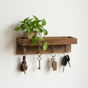 Puede incluir: Un llavero de madera con cuatro ganchos de metal y un pequeño estante, que contiene llaves y una planta en maceta. La madera tiene un acabado marrón cálido. Las llaves incluyen una llave vintage, llaves de coche y llaves con un llavero de cuero.