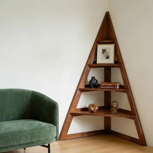 Puede incluir: Una estantería esquinera triangular de madera con cuatro niveles. Los estantes contienen una impresión abstracta enmarcada, un jarrón de cerámica negra, dos libros apilados titulados Form & Function y Modernism Redefined, un cubo de rompecabezas de madera y una botella de vidrio transparente.