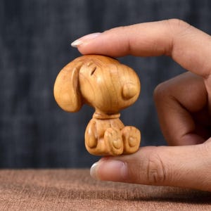 Può includere: Piccola statuina in legno di un cane dei cartoni animati con grandi orecchie e testa rotonda. La statuina è scolpita in legno chiaro e viene tenuta tra due dita. Il cane è seduto, con le zampe visibili.