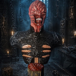 May include: A detailed bust of a humanoid creature with a red, textured, skinless-appearing head and exposed teeth. The figure wears a black, armored chest piece featuring horizontal, flesh-toned rib-like accents and sits atop a gold-toned puzzle box.