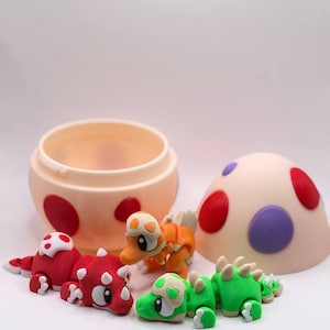 May include: A cream-coloured egg-shaped container with red and purple spots, accompanied by three small articulated dinosaur figures in red, orange, and green. Each dinosaur features a matching spotted shell piece on its head and back.