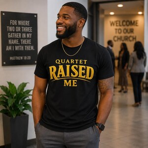 May include: A black short-sleeved t-shirt featuring the gold text QUARTET RAISED ME printed on the chest. The shirt is worn with a thin gold chain necklace and a black digital watch on the wrist, paired with dark grey trousers.