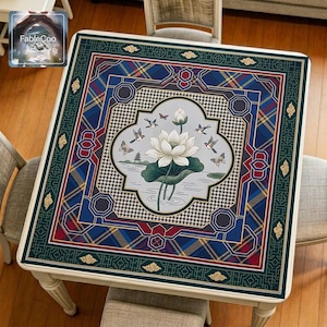 Asian-style mahjong mat displayed on a square home table, featuring an original oriental-inspired design with a central white lotus, broad lotus leaf, flying birds, butterflies, flowing water details, geometric medallion framing, Greek key accents, and richly layered ornamental borders in deep green, royal blue, burgundy red, gold, ivory, and soft gray tones.
