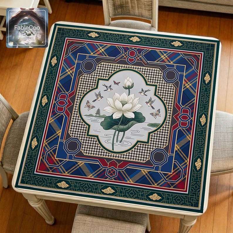 Asian-style mahjong mat displayed on a square home table, featuring an original oriental-inspired design with a central white lotus, broad lotus leaf, flying birds, butterflies, flowing water details, geometric medallion framing, Greek key accents, and richly layered ornamental borders in deep green, royal blue, burgundy red, gold, ivory, and soft gray tones.