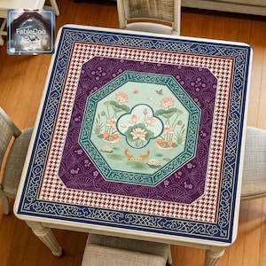 Asian-style mahjong mat displayed on a square home table, featuring an original oriental-inspired lotus pond design with blooming lotus flowers, lotus leaves, koi fish, small birds, butterflies, gentle water ripples, an octagonal window-style frame, fan-pattern detailing, houndstooth accents, and elegant decorative borders in navy blue, plum purple, teal green, soft mint, ivory, and gold tones.