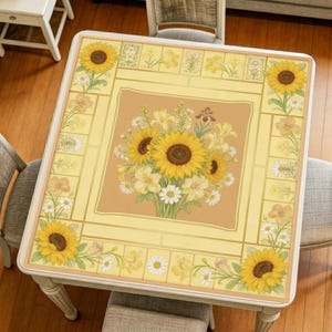 May include: A square, yellow tablecloth with a floral design featuring sunflowers and daisies. The tablecloth is on a white table, with a bouquet of sunflowers in the center. The table is surrounded by chairs.