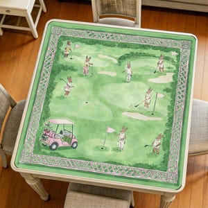 Pode incluir: Uma mesa quadrada com um design de campo de golfe verde com coelhos de desenho animado jogando golfe. A mesa tem uma moldura branca e um carrinho de golfe rosa. O design do campo de golfe é cercado por uma borda verde e rosa.