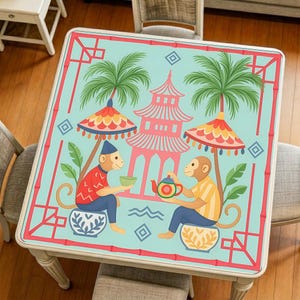 Op de afbeelding: Vierkante tafel met een kleurrijk, Aziatisch geïnspireerd ontwerp. De tafel heeft een lichtblauwe achtergrond met een roze pagode, palmbomen en apen. Decoratieve parasols en bamboe-achtige randen omlijsten het kunstwerk.