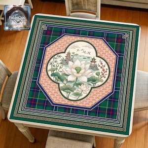 Asian-style mahjong mat displayed on a square home table, featuring an original oriental-inspired lotus pond scene with blooming lotus flowers, layered lotus leaves, traditional pavilions, flying birds, bamboo details, soft water elements, decorative window-style framing, Greek key borders, and tartan accents in deep green, navy blue, coral pink, gold, ivory, and muted natural tones.