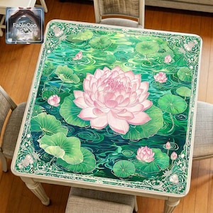 Asian-style mahjong mat displayed on a square home table, featuring an original lotus pond design with a large blooming pink lotus flower, layered lotus leaves, soft water ripples, lotus buds, and an elegant floral ornamental border in refreshing shades of emerald green, jade, deep teal, blush pink, and ivory.