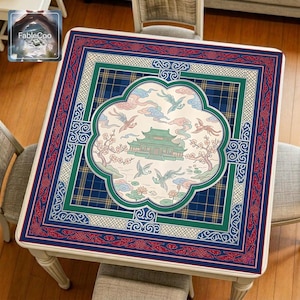 Asian palace-style mahjong mat displayed on a square home table, featuring an original oriental-inspired design with a central palace building, flying cranes, flowering branches, lotus details, soft cloud motifs, and layered decorative borders in deep blue, teal green, red, and ivory tones.