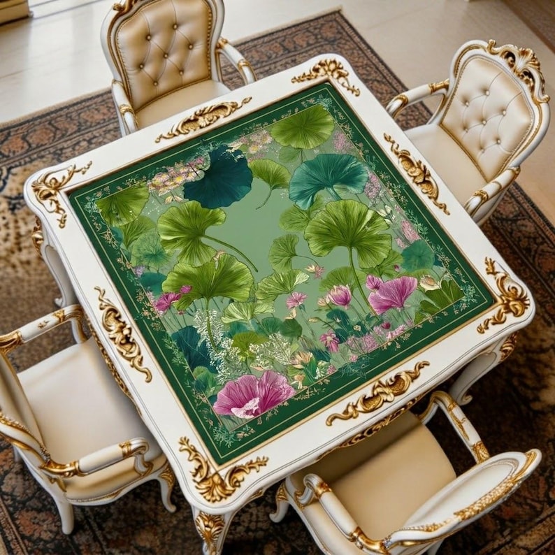 May include: A square white dining table with ornate gold trim and four matching chairs. The table's surface features a green floral design with large green leaves and pink flowers. The chairs have cream-colored upholstery and gold accents.
