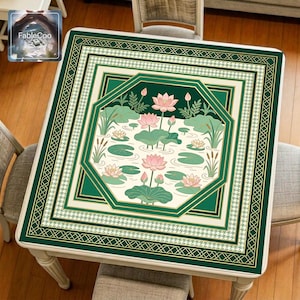 Asian-style mahjong mat displayed on a square home table, featuring an original oriental-inspired lotus pond design with blooming pink lotus flowers, broad lotus leaves, lily pads, reeds, flowing water lines, layered octagonal framing, and elegant gold borders in deep green, ivory, pale pink, soft jade, and muted gold tones.