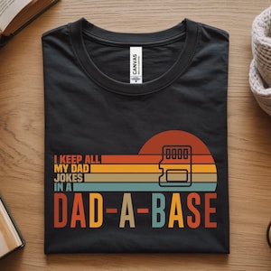 Może przedstawiać: Czarny t-shirt z okrągłym dekoltem, ozdobiony grafiką w stylu retro z poziomymi pasami w kolorze pomarańczowym, żółtym i niebieskim. Napis głosi I KEEP ALL MY DAD JOKES IN A DAD-A-BASE, obok znajduje się liniowa ikona karty pamięci SD.