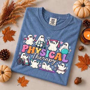 Op de afbeelding: Een blauw t-shirt met een afbeelding van witte geesten die fysiotherapie-oefeningen doen, waaronder met een rolstoel, rollator en oefenbal. De tekst "PHYSICAL Therapy" is gedrukt in grote, kleurrijke letters in retrostijl.