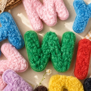 Peut inclure: Un assortiment de lettres en peluche colorées, dans diverses teintes, dont le rose, le bleu, le vert, le rouge, le jaune et le violet. Les lettres sont faites d'une matière douce et texturée, disposées sur une surface claire.