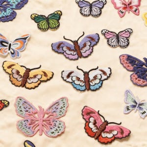 Pode incluir: Uma coleção de emblemas bordados de borboletas em várias cores, incluindo rosa, amarelo, azul, roxo e verde. Cada emblema apresenta um trabalho de fio detalhado que retrata asas com padrões distintos, corpos escuros e antenas sobre um fundo creme.