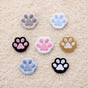 Peut inclure: Plusieurs écussons brodés en forme de pattes de chat, de couleurs variées : rose, bleu, noir et or. Les écussons sont posés sur une surface texturée blanc cassé. Parfaits pour décorer vêtements et accessoires.