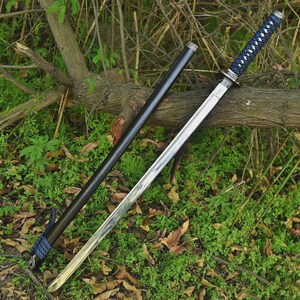 Op de afbeelding: Een katana-zwaard met een gepolijst zilveren lemmet en een handvat omwikkeld met blauw en wit koord. Het zwaard wordt vergezeld door een gladde, zwarte schede met een bijpassend blauw en wit koordaccent bij de basis, rustend tegen een tak.
