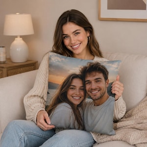 Può includere: Un cuscino rettangolare con una fotografia di una coppia sorridente sullo sfondo di un tramonto. Il cuscino è tenuto da una persona che indossa un maglione color crema e jeans blu, seduta su un divano beige.