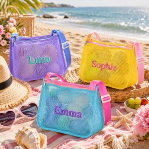 Puede incluir: Tres bolsas de playa de malla en morado, amarillo y azul, cada una bordada con un nombre: Luna, Sophie y Emma. Las bolsas tienen correas y cremalleras a juego. Las bolsas están en una playa de arena.