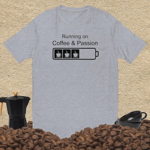 May include: A heather gray short-sleeve t-shirt featuring the black text Running on Coffee & Passion above a battery icon graphic containing three coffee cup symbols. The design is centered on the chest of the shirt, which has a crew neckline.
